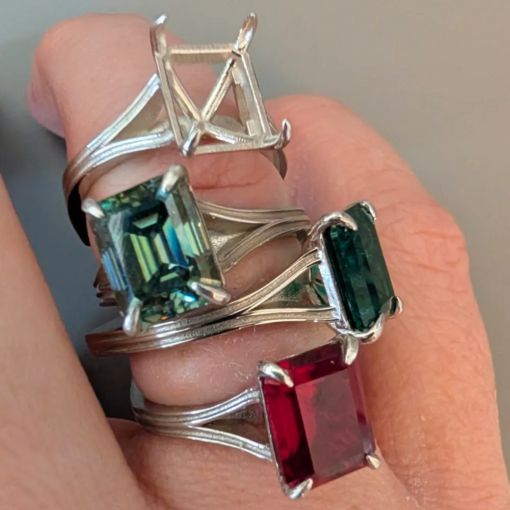 Four rings stacked on a hand showing emerald-cut green sapphires and a ruby in split-shank settings